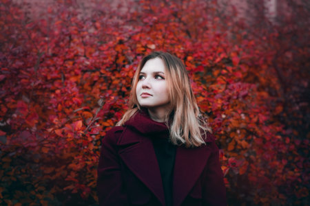 Portrait of a beautiful blonde woman in a burgundy coat against the background of red leaves of...