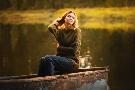 A beautiful blonde woman is sitting on the edge of an old fishing boat floating in a lake in the forest and enjoying the beauty of nature. Travel and recreation.の写真素材
