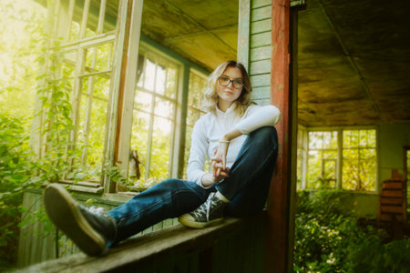 A cute beautiful teenager with blonde hair and jeans is sitting on the windowsill of a wooden abandoned house on a sunny summer day.の写真素材
