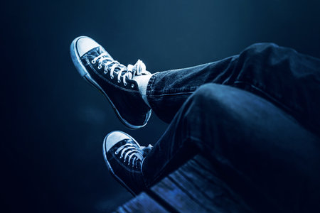 Legs in sneakers and blue jeans of a human sitting on a wooden pier above the dark water of the lake. Lifestyle and recreation by the water.の写真素材