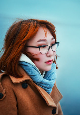 Portrait of a beautiful ginger Asian woman with glasses, wearing a blue scarf and a beige coat against the background of a cold sea on a winter day. The romance of travel. cold season.の写真素材