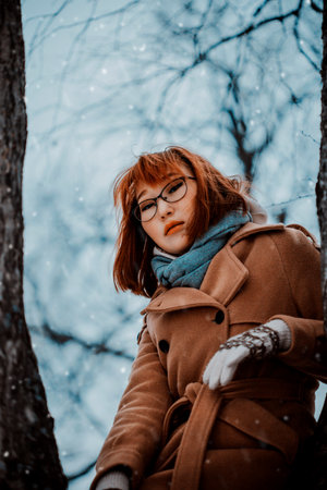 Portrait of a beautiful ginger Asian woman in warm clothes, sitting leaning on a tree trunk on a snowy winter day. January weather.の写真素材