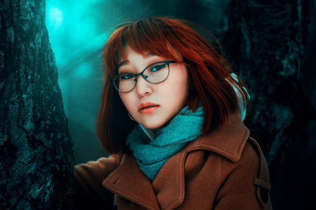 A close-up portrait of a beautiful Asian red-haired woman in warm clothes in a dark forest near tree...