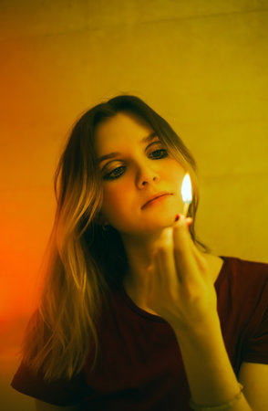 Portrait of a beautiful blonde woman who looks at a brightly burning match, which she holds in her hand in the semi-darkness. house hold items.の写真素材