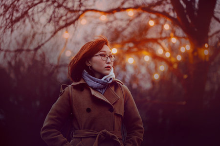 Portrait of a beautiful Asian woman in a coat standing in a park near a tree with a glowing garland on a dark autumn evening. decorations.の写真素材