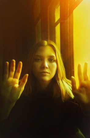 Portrait of a beautiful young blonde woman who is on the other side of an old window glass and touches its surface with her hands, illuminated by an otherworldly yellow light.の写真素材