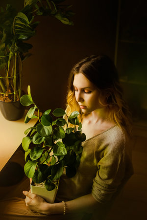 Portrait of a beautiful young blonde in beige clothes, who is sitting with a potted green flower in her hands at home. love of plants. Home gardening and plant breeding. Ecology.の写真素材