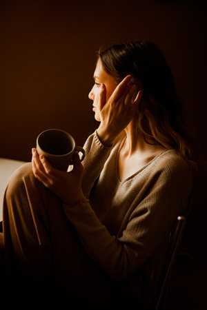 A sweet, beautiful young blonde in beige clothes is sitting in the kitchen alone in the morning, holding an empty mug in which tea has run out. lifestyle. Loneliness.の写真素材