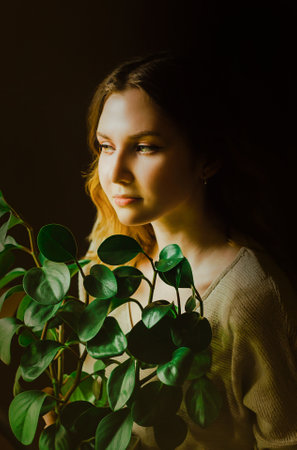 Portrait of a beautiful blonde young woman in a beige sweater, who is sitting close to a green plant that she has grown. Home gardening and love of plants. Ecology and nature protection.の写真素材