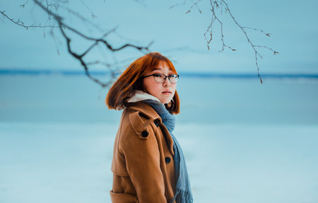 Portrait of a beautiful red-haired young Asian woman in a beige coat, who walks near the frozen sea in winter in the park. Travel in the cold season. North.の写真素材