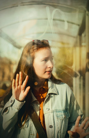 Portrait of a beautiful young red-haired woman in casual clothes, who stands behind the glass of a bus stop and waits for the bus on a sunny summer day.の写真素材