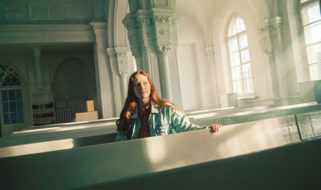 A beautiful young red-haired woman is sitting on old pews inside a Protestant church, illuminated by the radiance of the sun's rays and divine light. Faith and religion.の写真素材
