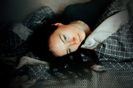 Portrait of a beautiful young woman lying on a soft bed with a patterned plaid on a dark sad...