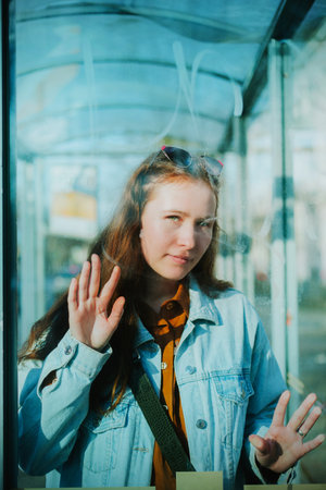 Portrait of a beautiful young woman in a denim jacket who is waiting for a bus at a bus stop. Portrait through transparent glass.の写真素材