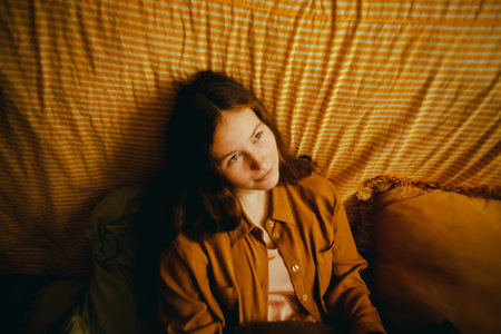 Portrait of a beautiful young woman in a brown shirt sitting on a bed among soft pillows in the early morning. Lazy morning.の写真素材