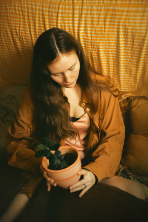 A woman sitting on a bed inside a home while holding a potted plant. Relaxation and the natural beauty of indoor gardening, illustrating how plants can enhance the ambiance of a living space.の写真素材