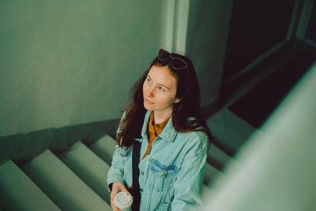 An young female student is seen ascending a staircase with a cup of coffee in her possession. She appears to be returning from a brief interval, all set to embark upon her next task. businesswoman.の写真素材
