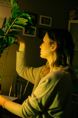 A young blonde woman in casual clothes taking care of her houseplant at home in the evening....