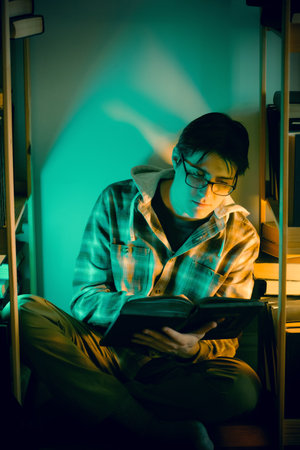 A young man in a checkered shirt and glasses is sitting among bookshelves late at night and reading a book. Studying, self-education, the pursuit of knowledge and reading alone in a library.の写真素材