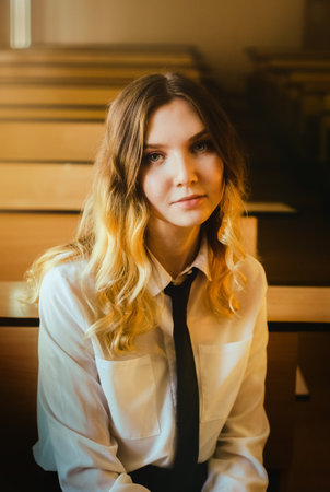 A beautiful blonde schoolgirl dressed in a white shirt and black tie is sitting alone at a school desk in a sunny classroom on a bright day. Educational environment, discipline and loneliness.の写真素材