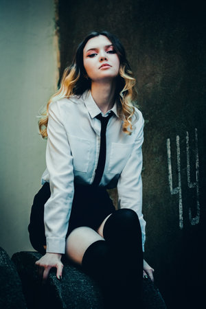 A beautiful young woman in a school uniform, sitting on a concrete circle near an old concrete wall. Grunge and youth.の写真素材