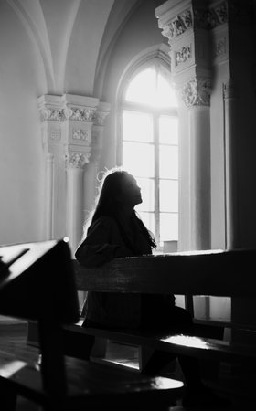 The black-white image depicts a young woman sitting on a bench in an old church, illuminated by bright rays of sunlight. Faith, spirituality, and introspection.の写真素材