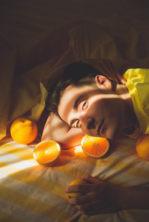 A young handsome man in a yellow t-shirt lying on a bed with oranges, illuminated by warm sunlight on a summer day. Relaxation, health, and enjoying the simple pleasures of life. fruits.の写真素材