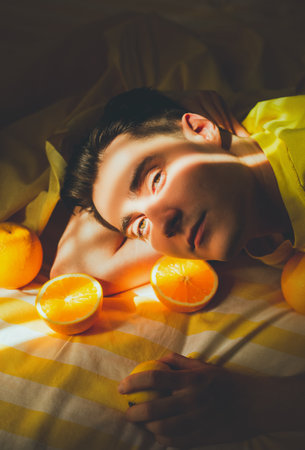 A portrait of a young handsome man, lying on a striped yellow bed amidst oranges, illuminated by warm rays of the summer sun. Youth, beauty, relaxation, and the vibrant energy of summer. Season.の写真素材