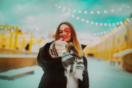 A young, joyous woman stands at a winter fair, holding a red Christmas candy cane and smiling at the camera. The celebration of the New Year, evoking themes of holiday cheer, festivity and season.の写真素材