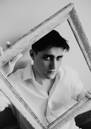 The black-white photograph captures a young handsome man in a white shirt sitting next to a white wall, holding an empty picture frame in his hands. The connection between the artist and his craft.の写真素材