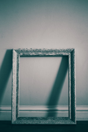 A dimly lit image of a white and beautiful empty frame standing against an empty wall in a gallery. A preparation for an upcoming exhibition, with no one present in the room. Emptiness.の写真素材