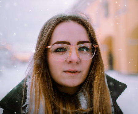A portrait of a beautiful girl wearing glasses and warm clothing on a winter walk during a snowy day. Winter, cold weather, and the beauty of snowfall.の写真素材