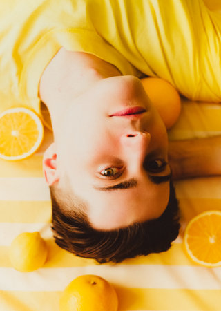A portrait of a young man lying amidst citrus fruits on a striped yellow bed, illuminated by warm rays of summer sunshine. oranges. Youth, vitality, and the pleasure of enjoying the fruits of life.の写真素材