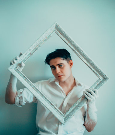 A man with his hands painted in white, holding an empty painting frame. An art gallery setting, with the man preparing for an exhibition. Creativity, art, and the process of preparing for a showcase.の写真素材