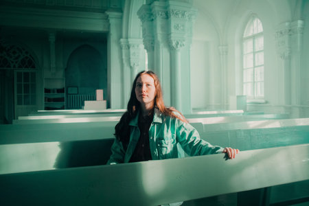 A beautiful young ginger woman is sitting thoughtfully on a bench in a Lutheran church, wearing a denim jacket. The religion, introspection, and contemplation. Faith.の写真素材
