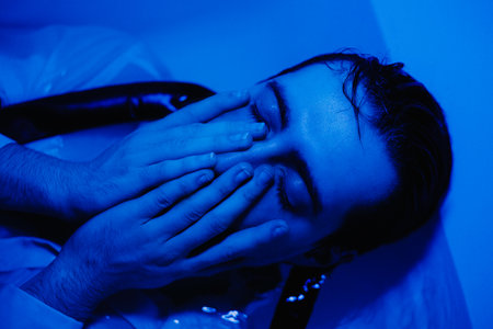 A sad, wet man in a shirt lies with his eyes closed in a bath filled with water, illuminated in the dark by blue light and touches his face with his hands. Depression and melancholy. Burnout at work.の写真素材