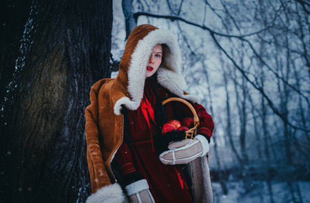 Portrait of a young woman in folklore clothes, fur sheepskin coat and mittens, standing in the middle of a winter forest with a basket of apples in her hands. Slavic fairy tale and fantasy.の写真素材