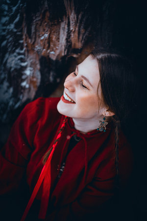 A cute girl with pigtails in a scarlet Slavic dress sits near a tree in the snowy winter in the forest and smiles. Folklore and fairy tale. Traditions.の写真素材