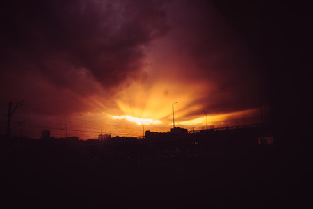 The urban landscape at night. The setting sun casts bright rays through the clouds, illuminating the silhouette of the city skyline and the bridge.の写真素材