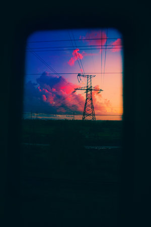 From the window of the train, I saw a beautiful landscape as the sun rose. There was an electric grid tower in a field, with long wires stretching from it. The clouds in the sky had turned pink.の写真素材