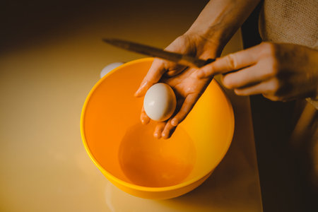 A woman is preparing to crack an egg using a knife, which she holds above a yellow bowl. This is a scene from a cooking process at home.の写真素材