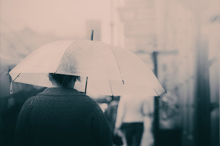 A lonely girl walks down a city street on a foggy day, holding an umbrella that shields her from the light rain. The monochrome colors of the scene add a sense of sadness and melancholy.の写真素材