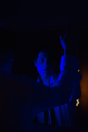 A young man stands in a semi-dark room, illuminated by a blue light, looking at his reflection in a mirror.の写真素材