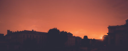 The setting sun casts a beautiful glow over the dark silhouettes of the city rooftops, turning the sky a warm orange color.の写真素材
