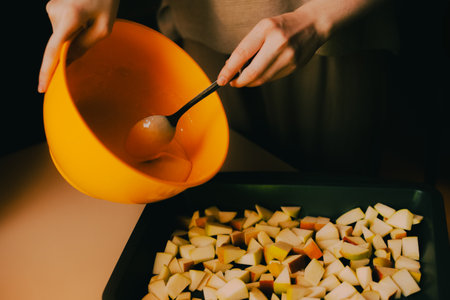 A woman in casual attire is pouring batter from a bowl onto a baking tray filled with deliciously sliced apples, preparing a homemade apple cake.の写真素材