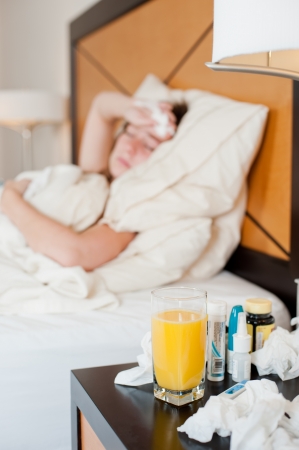 A sick young lady in bed out of focus in the background, with focus on the orange juice on the bedside table, with some medicine visible の写真素材