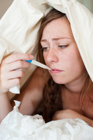 A sick young lady in bed, using a thermometer to check her temperature for fever の写真素材