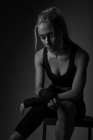 Serious young professional female kickboxer wearing black sportswear and bandages on hands, posing in dark atmosphere. Fighter having rest after trainingの写真素材