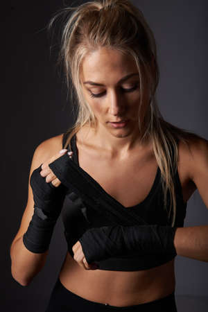 Serious young professional female kickboxer wearing black sportswear and bandages on hands, posing in dark atmosphere. Fighter having rest after trainingの写真素材