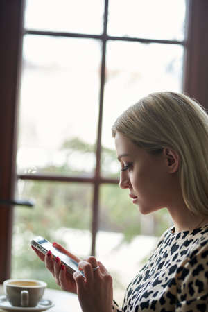 Image of young woman reading sms by phone in cafe with cup of coffeeの写真素材